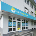 Eingangsbereich eines sportwetten.de-Wettbüros. Über dem Eingang befindet  
sich ein türkisfarbenes Schild mit dem sportwetten.de-Logo, in den Farben Weiß und Gelb.  
Große Fenster und eine Glastür sorgen für ein einladendes, modernes Erscheinungsbild.