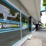 Schaufenster eines sportwetten.de-Wettbüros mit großen Glasfenstern,  
auf denen in fetten Lettern das Logo „sportwetten.de“ zu  
lesen ist, mit Blick auf eine ruhige Straße und grüne Bäume.