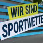 Das Bild zeigt ein sich überlappendes Folienmotiv auf einem  
Schaufenster mit türkisfarbenen Streifen. Darauf steht in dunkelblauen  
Buchstaben auf Gelb und Weiß „Wir sind Sportwetten“.