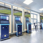 Ein modernes sportwetten.de-Wettbüro mit vier aufgereihten Wettterminals.  
Auf großen Bildschirmen wird ein Fußballspiel gezeigt, und in der  
Nähe steht ein Stehtisch mit Hockern.
