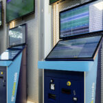 Eine Reihe von blauen sportwetten.de-Wettterminals mit großen  
Bildschirmen vor einem modernen Metallgitter-Hintergrund.