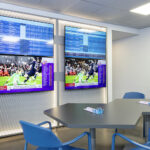 Ein sportwetten.de-Wettbüro mit großen Bildschirmen, auf denen Sportereignisse  
und Quoten angezeigt werden. Ein Tisch mit blauen Stühlen und  
Wettscheinen schafft eine konzentrierte Atmosphäre.