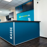 Innenansicht eines sportwetten.de-Wettbüros mit Blick auf die blaue Kasse mit  
der Aufschrift „KASSE“. Hinter der Kasse ist eine türkisfarbene Wand zu sehen mit dem  
sportwetten.de-Logo in der Mitte. Darüber hängen zwei Monitore die Quoten anzeigen.